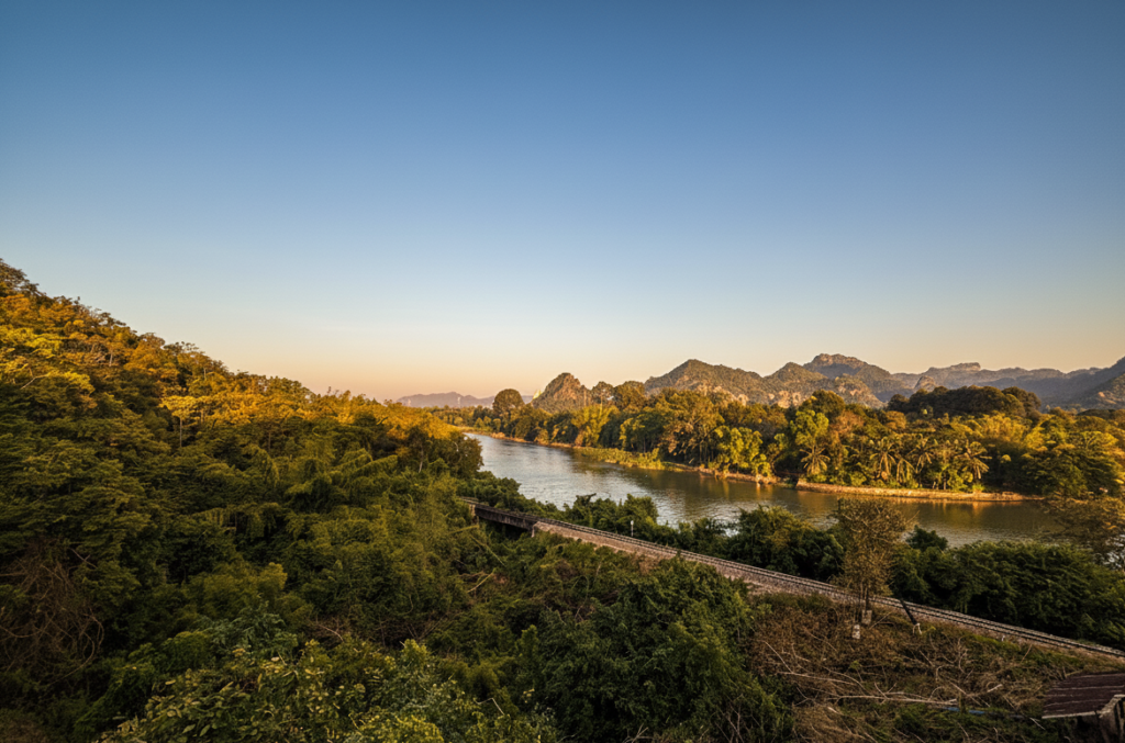 A high-angle landscape shot of the historic Thai-Burma Railway tracks following the curve of the Khwae Noi River, surrounded by dense tropical forest and limestone hills.